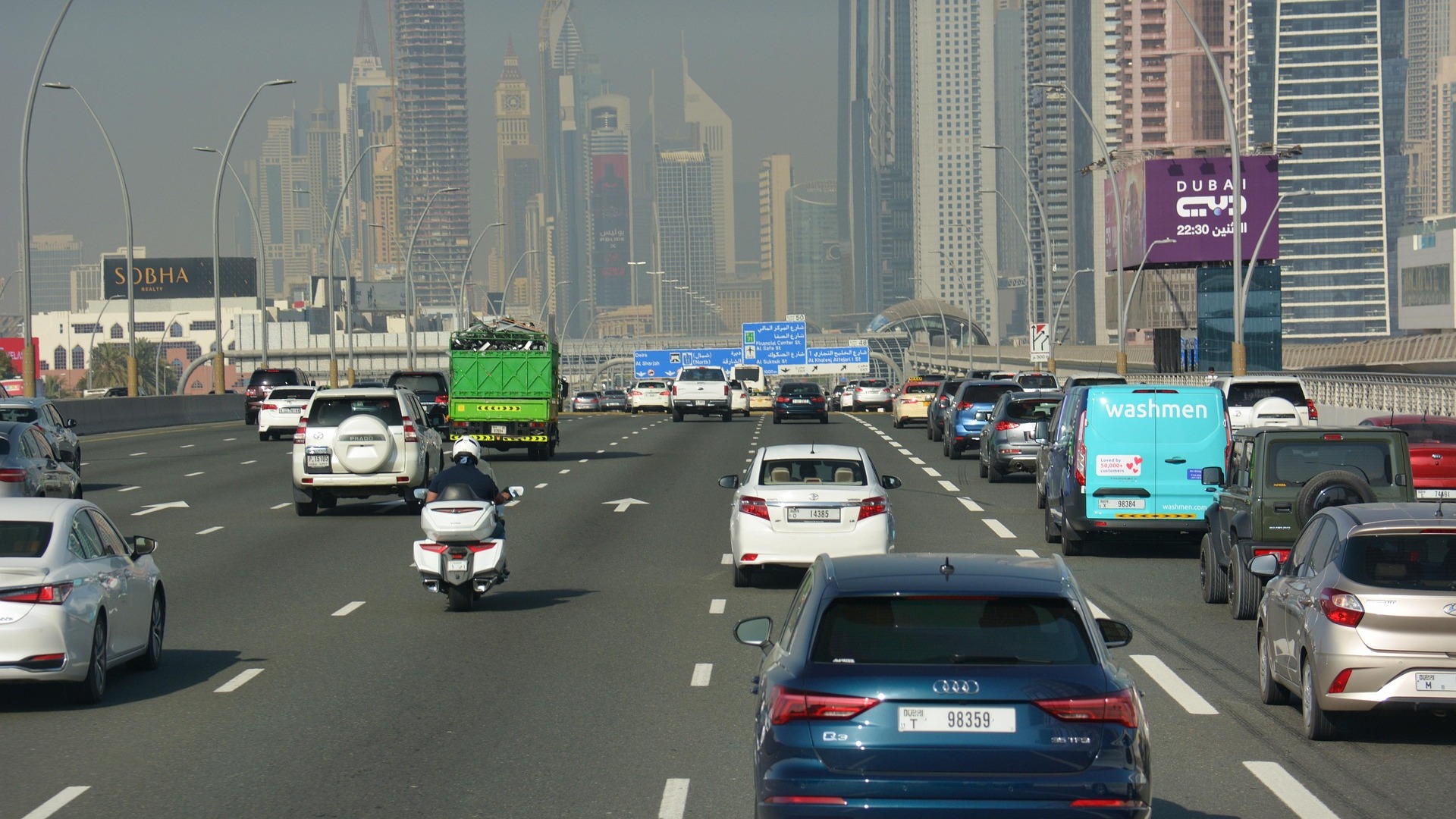 Heavy traffic filling every lane of a multi-lane urban expressway, with a hazy city skyline behind it.