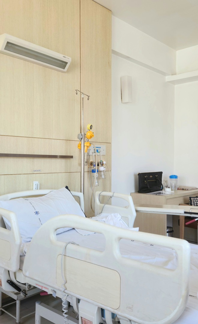 A quiet hospital room with made-up beds, a drip stand and a chair by the window — a recovery ward, not an operating theatre.