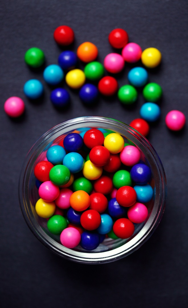 A glass bowl of brightly colored gumballs with more scattered on the surface beside it. Red is the most common color.