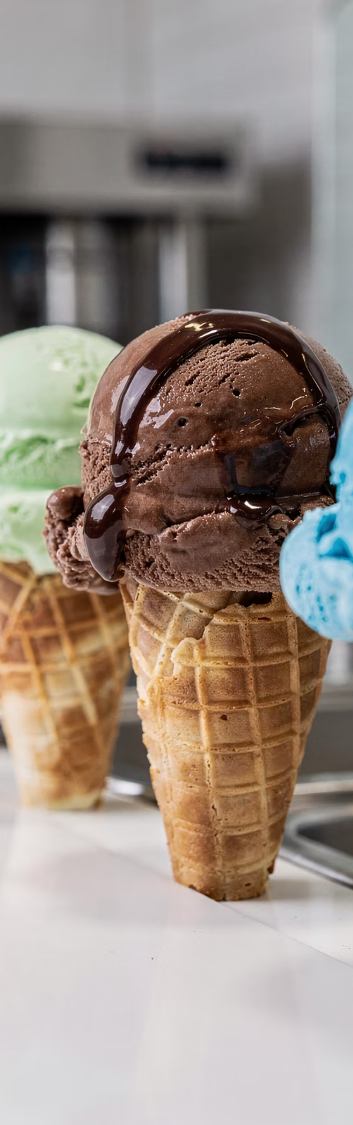 Three ice cream cones in a row, a chocolate scoop with fudge sauce in front.